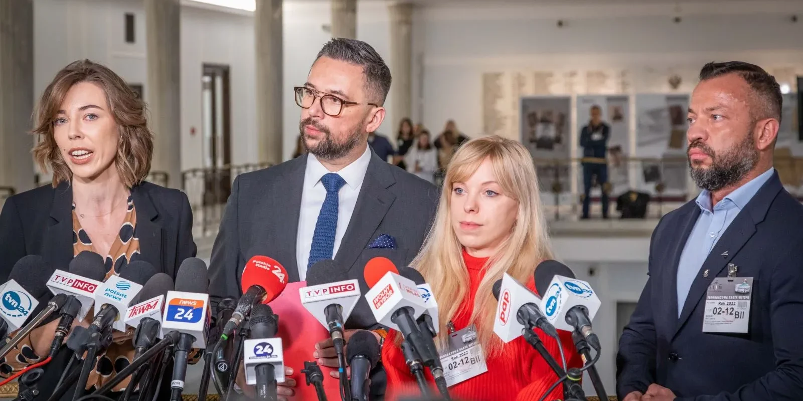 Cztery osoby stojące przed mikrofonami na konferencji prasowej w jasnym wnętrzu.