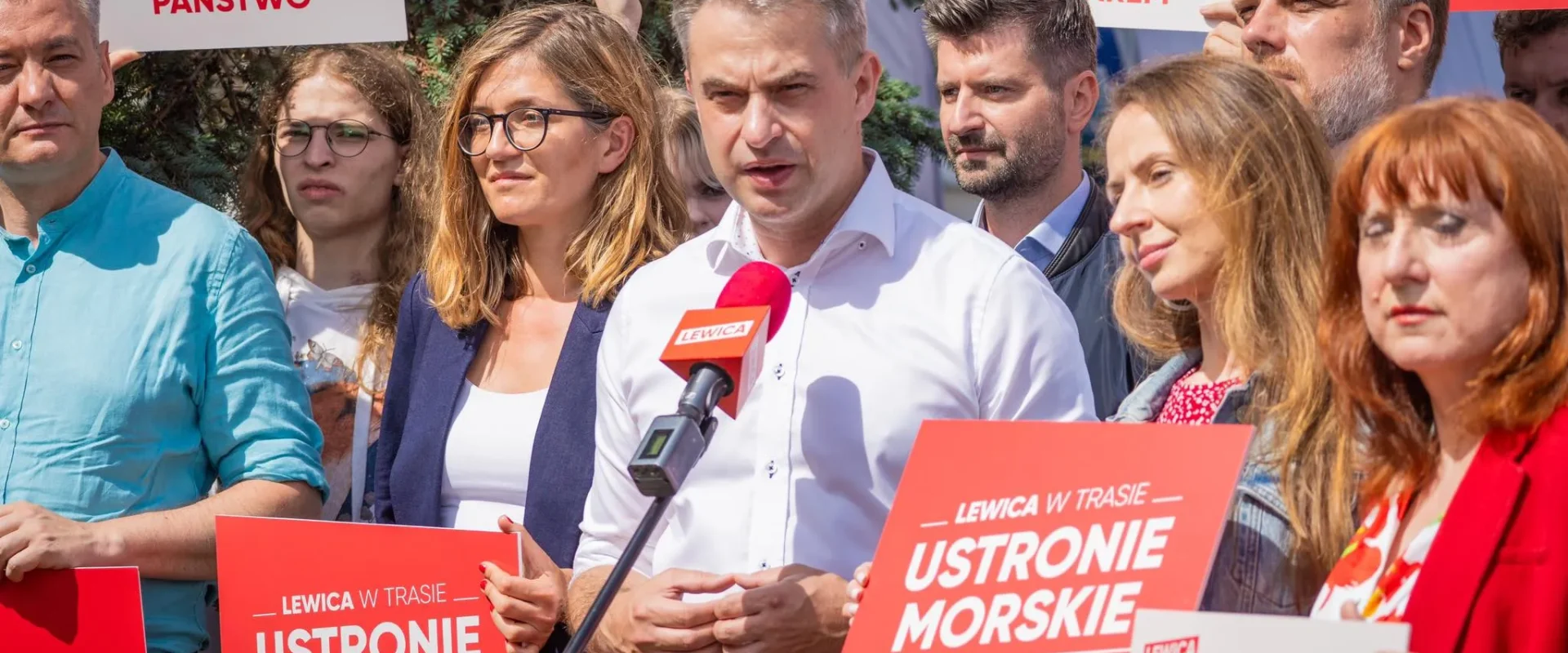 Grupa ludzi na wiecu politycznym trzyma czerwone plakaty Lewica w trasie i przemawia do mikrofonu.