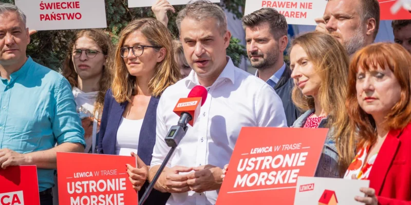 Grupa ludzi na wiecu politycznym trzyma czerwone plakaty Lewica w trasie i przemawia do mikrofonu.