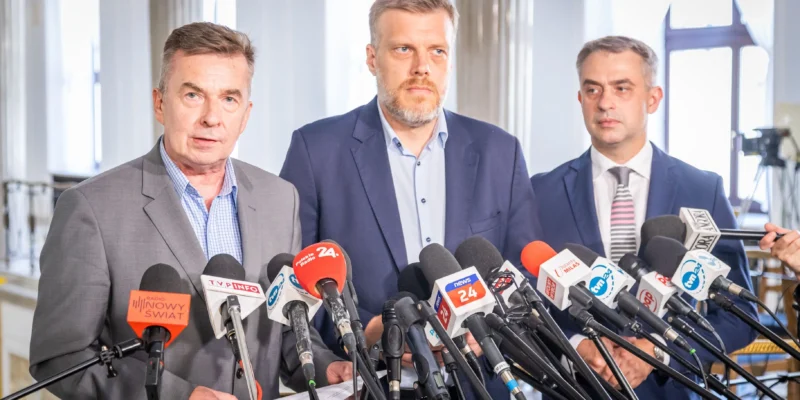 Trzech mężczyzn w garniturach stoi za mikrofonami na konferencji prasowej w jasnym wnętrzu.