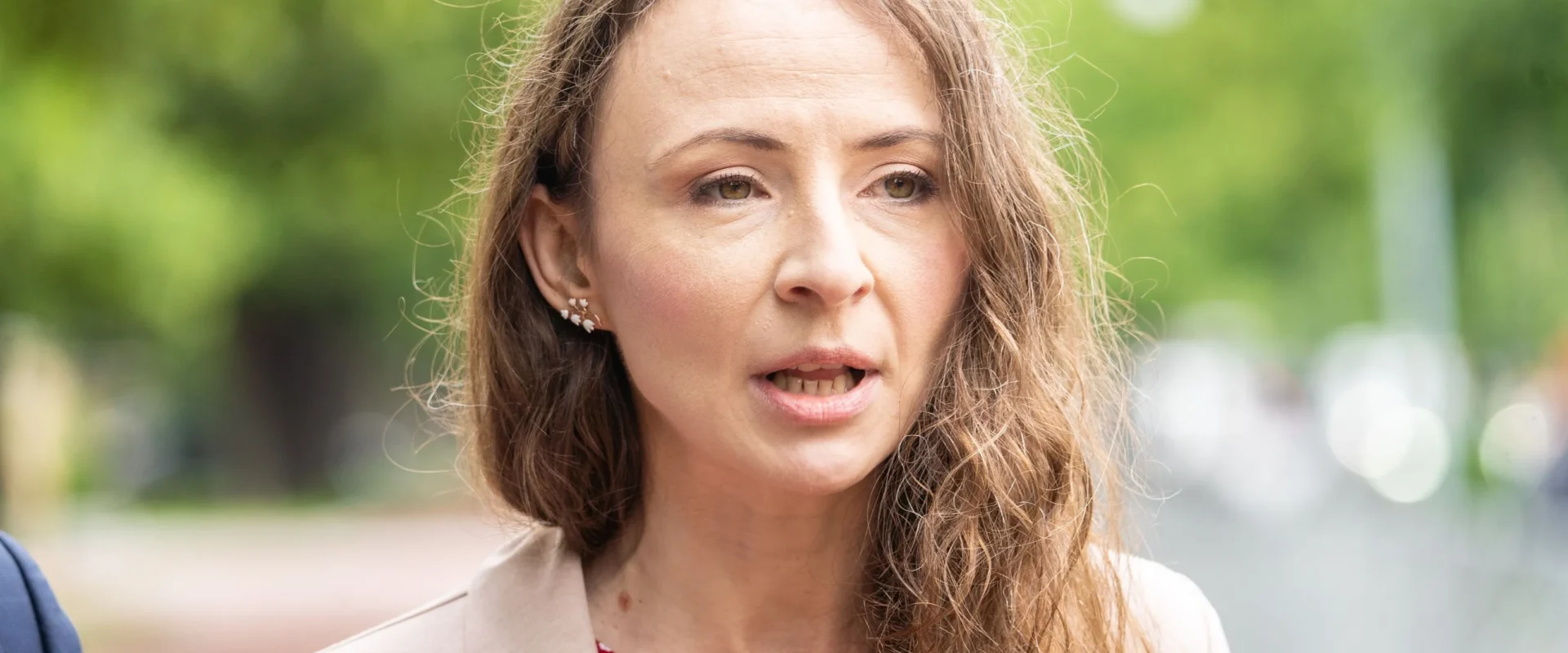 Kobieta o długich, falowanych włosach, ubrana w beżowy żakiet i czerwoną bluzkę w drobny wzór, przemawia na zewnątrz na tle zielonego, rozmytego tła.