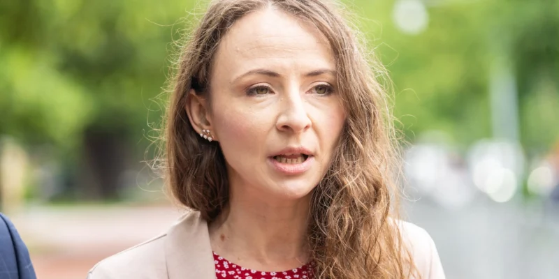 Kobieta o długich, falowanych włosach, ubrana w beżowy żakiet i czerwoną bluzkę w drobny wzór, przemawia na zewnątrz na tle zielonego, rozmytego tła.