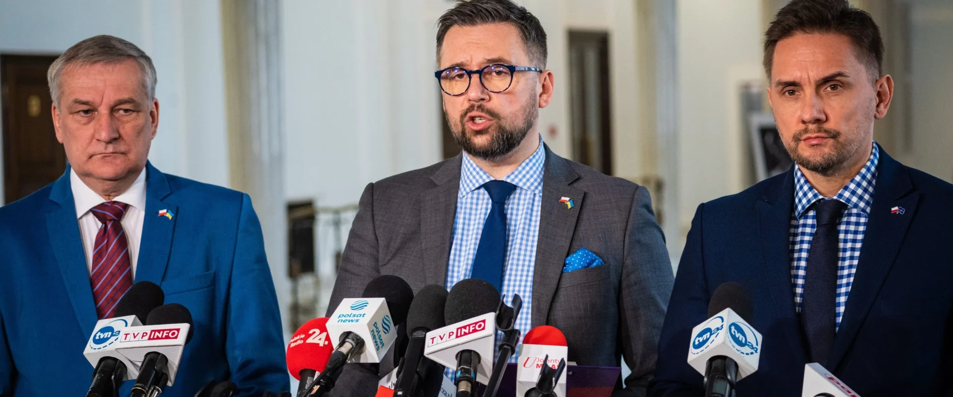 Trzech mężczyzn w garniturach stoi za mikrofonami podczas konferencji prasowej w eleganckim wnętrzu.