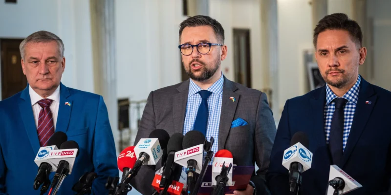 Trzech mężczyzn w garniturach stoi za mikrofonami podczas konferencji prasowej w eleganckim wnętrzu.