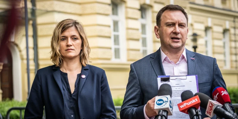 Dwóch polityków lewicy stoi przed budynkiem podczas konferencji prasowej; mężczyzna po prawej trzyma dokument i przemawia do mikrofonów, obok stoi kobieta.