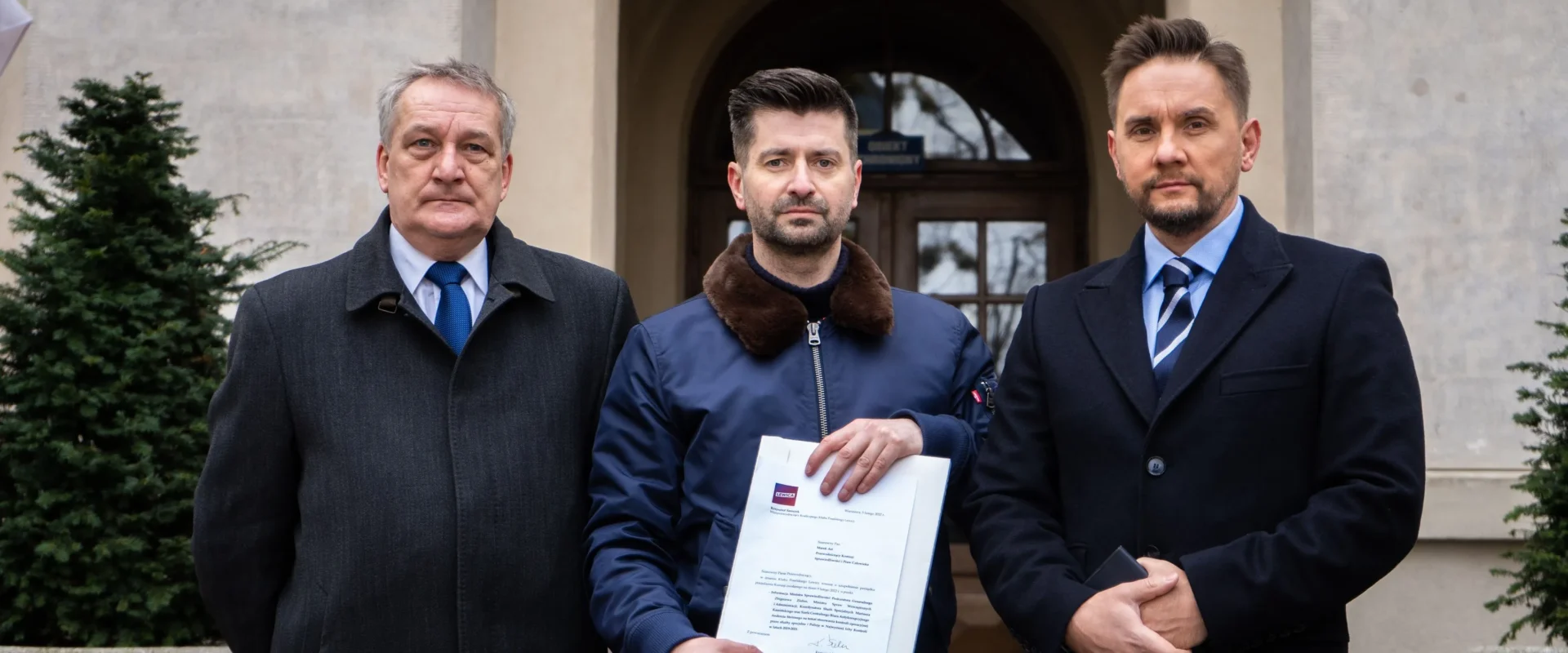 Trzech mężczyzn stoi przed wejściem do kamiennego budynku, a środkowy trzyma w dłoni dokumenty.