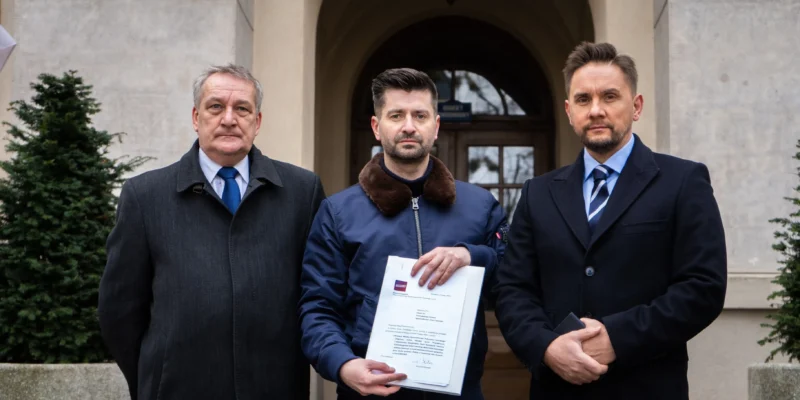 Trzech mężczyzn stoi przed wejściem do kamiennego budynku, a środkowy trzyma w dłoni dokumenty.