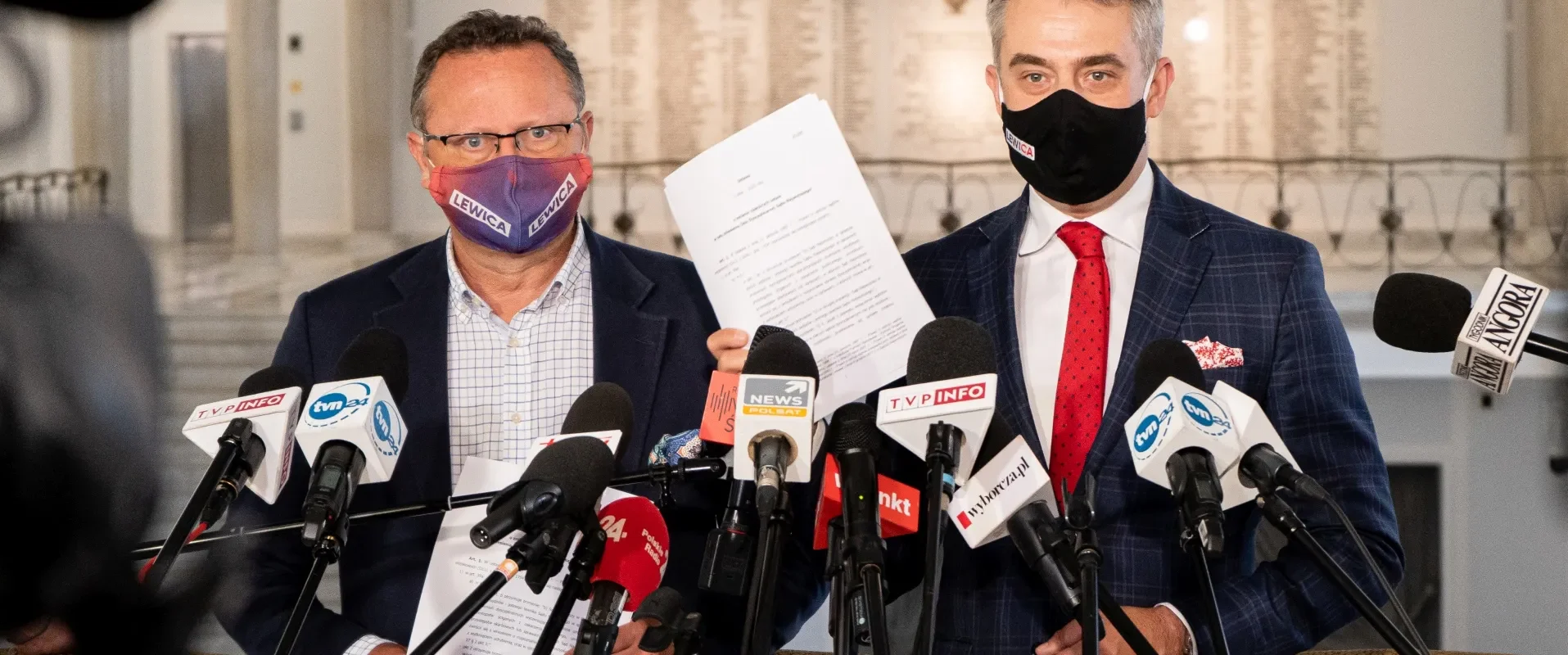 Dwóch mężczyzn w garniturach na konferencji prasowej stoi przed licznymi mikrofonami, trzymając dokumenty; jeden ma fioletową maseczkę z napisem Lewica, a drugi czarną maseczkę.
