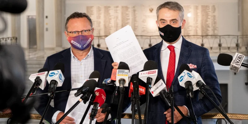 Dwóch mężczyzn w garniturach na konferencji prasowej stoi przed licznymi mikrofonami, trzymając dokumenty; jeden ma fioletową maseczkę z napisem Lewica, a drugi czarną maseczkę.