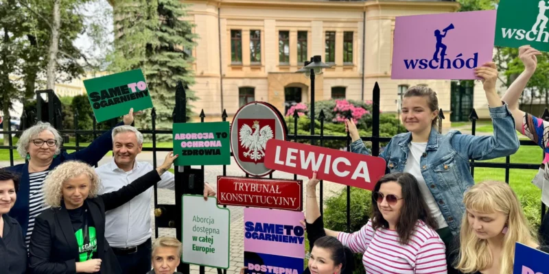 Grupa osób stojąca przy ogrodzeniu przed budynkiem trzyma kolorowe plakaty popierające Lewicę i europejską inicjatywę obywatelską „My voice my choice”.