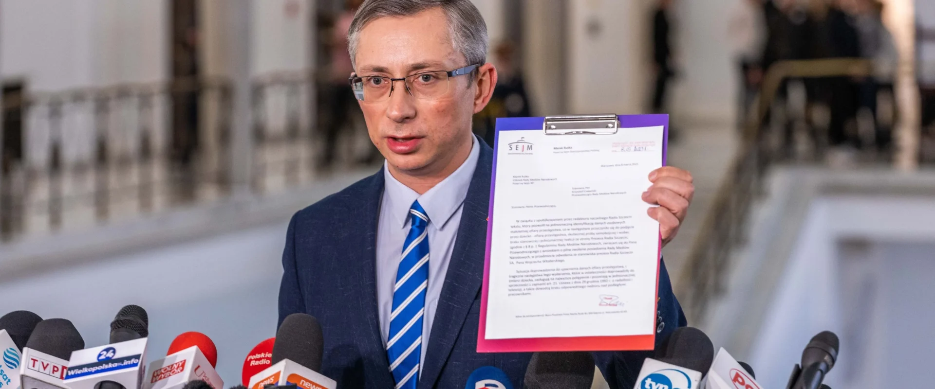 Polityk trzymający dokument podczas konferencji prasowej otoczony mikrofonami i dziennikarzami.