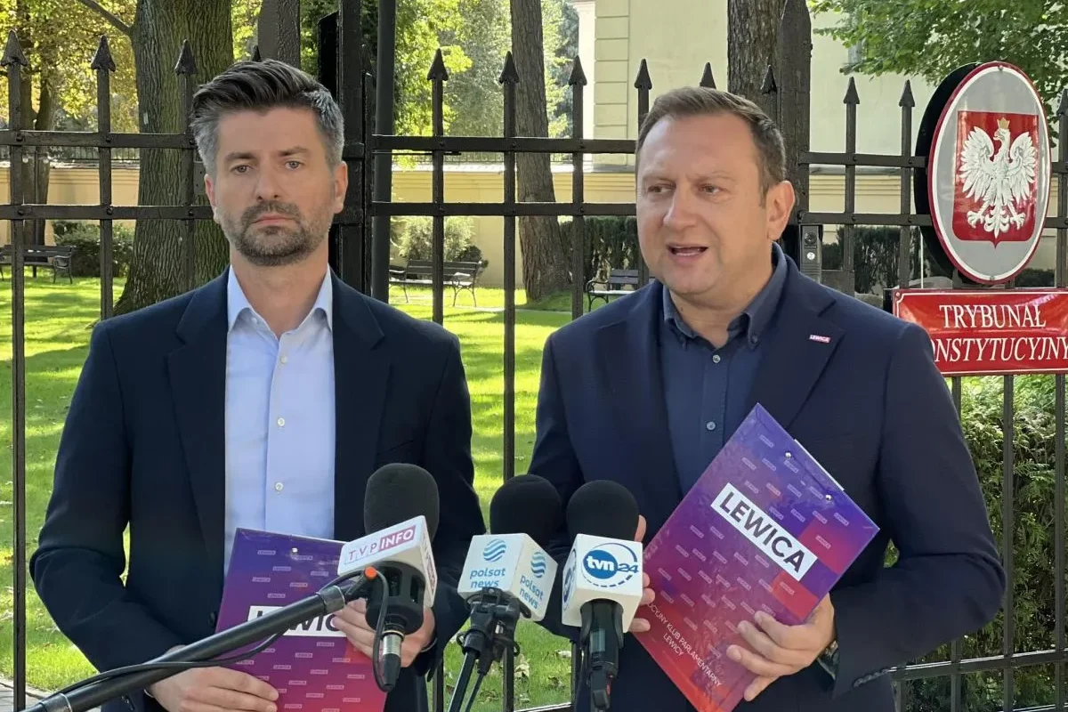 Dwóch mężczyzn w garniturach stoi przed bramą z godłem Polski i napisem Trybunał Konstytucyjny, trzymając fioletowe foldery z napisem Lewica i udzielając wywiadu mediom.