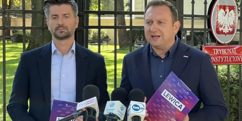 Dwóch mężczyzn w garniturach stoi przed bramą z godłem Polski i napisem Trybunał Konstytucyjny, trzymając fioletowe foldery z napisem Lewica i udzielając wywiadu mediom.