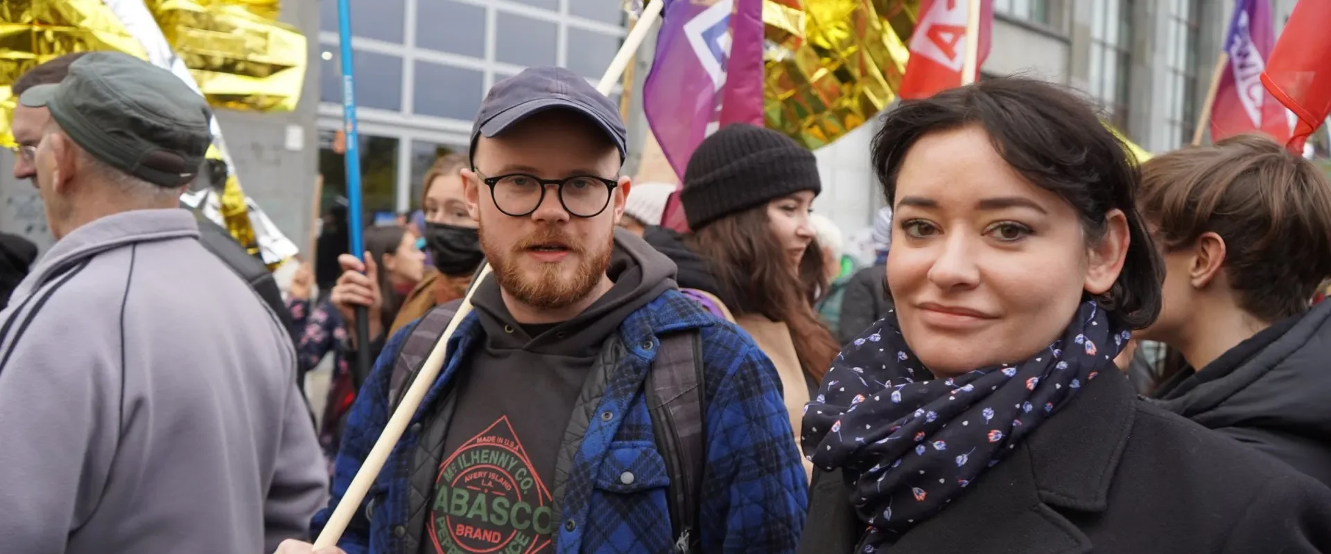 Uczestnicy marszu solidarności z migrantami na ulicy, w pierwszym planie mężczyzna z okularami i czapką oraz kobieta w czarnym płaszu i wzorzystym szaliku, otoczeni kolorowymi flagami.