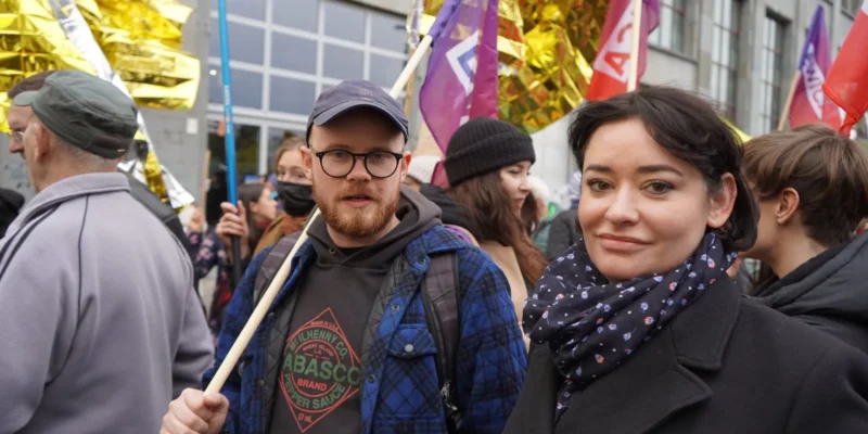 Uczestnicy marszu solidarności z migrantami na ulicy, w pierwszym planie mężczyzna z okularami i czapką oraz kobieta w czarnym płaszu i wzorzystym szaliku, otoczeni kolorowymi flagami.