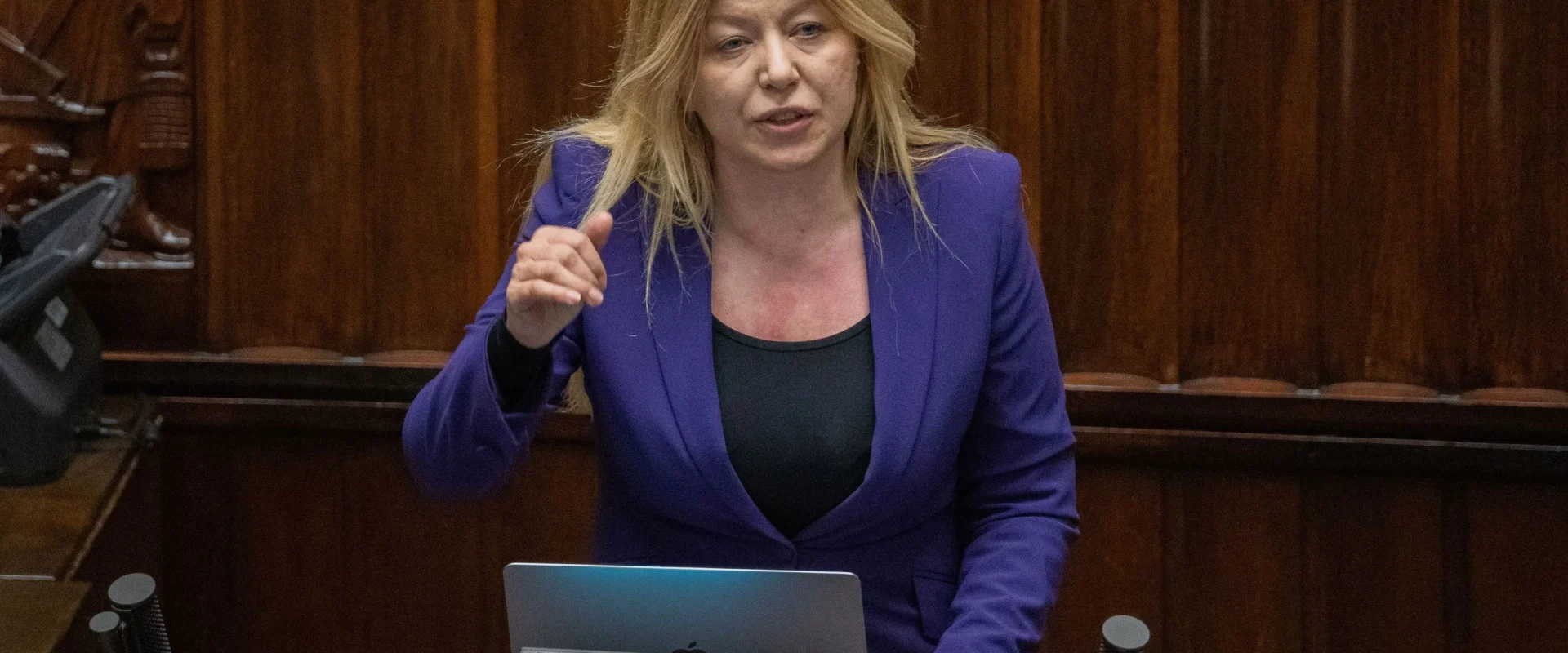 Kobieta o blond włosach w fioletowym żakiecie stoi przy mównicy z laptopem podczas przemówienia w sali plenarnej.