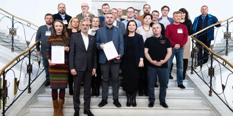 Grupa osób pozująca do zdjęcia na schodach, trzymająca dokumenty, w tle elegancki budynek.