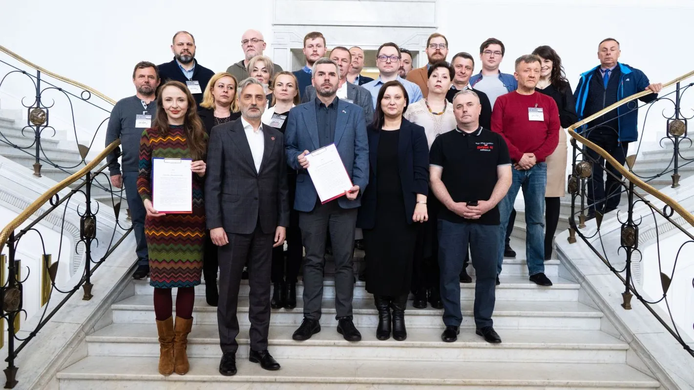 Grupa osób pozująca do zdjęcia na schodach, trzymająca dokumenty, w tle elegancki budynek.
