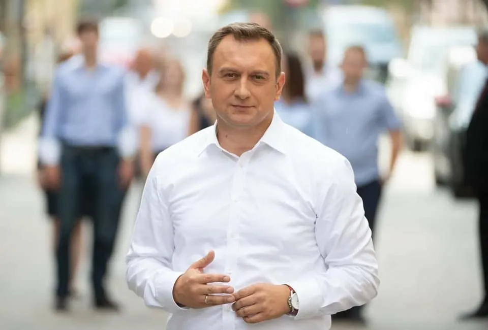 Mężczyzna w białej koszuli stoi na ulicy, a w tle widać rozmytą grupę ludzi.