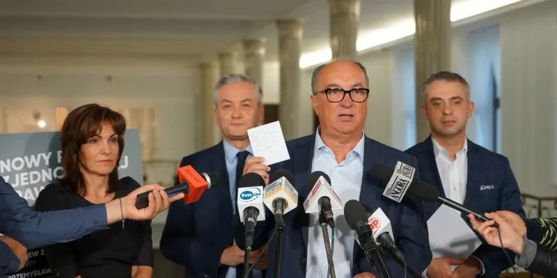 Grupa polityków na konferencji prasowej stoi za zestawem mikrofonów, a jeden z nich trzyma krótką notatkę.