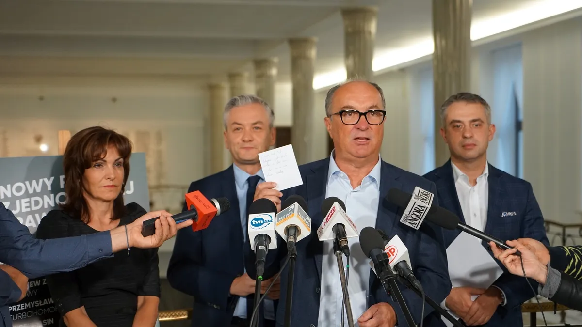 Grupa polityków na konferencji prasowej stoi za zestawem mikrofonów, a jeden z nich trzyma krótką notatkę.