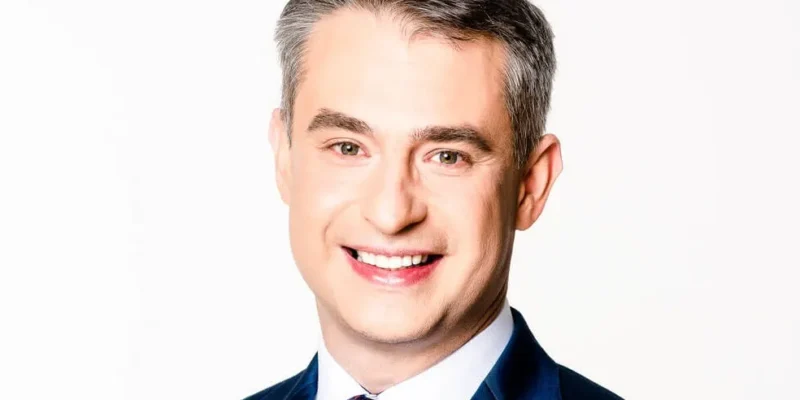 Uśmiechnięty mężczyzna w granatowym garniturze i czerwono-niebieskim krawacie na białym tle.