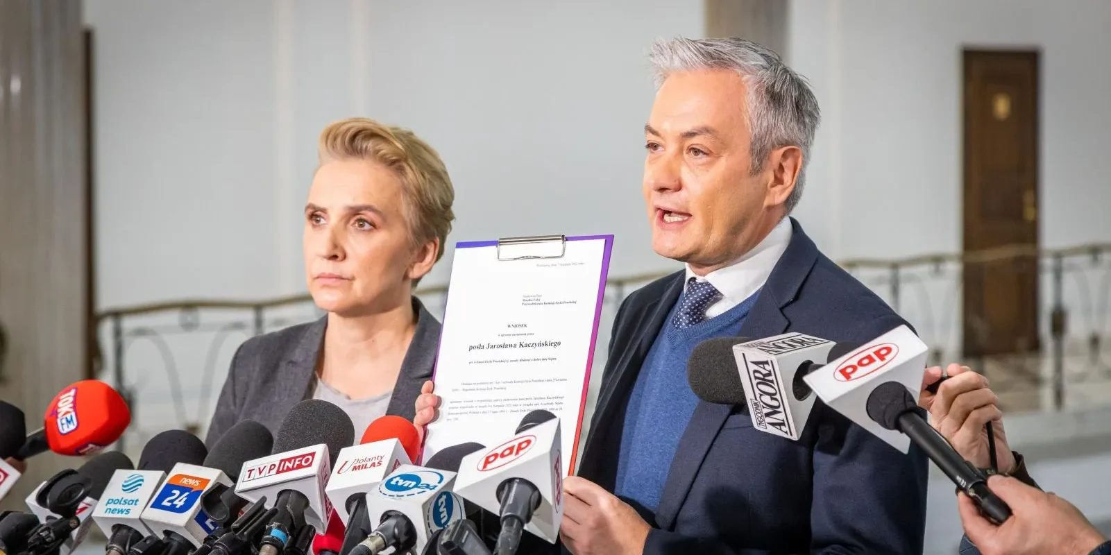 Na zdjęciu widoczna jest para polityków podczas konferencji prasowej, przy zebranych mikrofonach, z jednym z nich trzymającym dokument.