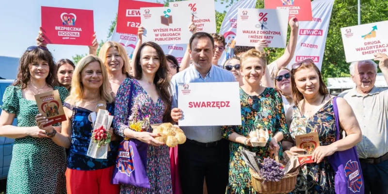Grupa osób na zewnątrz podczas wydarzenia politycznego trzyma banery Lewicy i kartki z hasłami Bezpieczna Rodzina, a w centralnej części ktoś trzyma plakat z napisem Swarzędz; scena ilustruje obawy przed rekordową inflacją zagrażającą rodzinom.