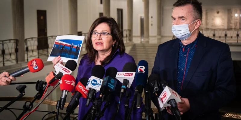 Na zdjęciu widać kobietę prezentującą zdjęcie podczas konferencji prasowej, obok której stoi mężczyzna, a w tle zbiera się grupa dziennikarzy z mikrofonami.