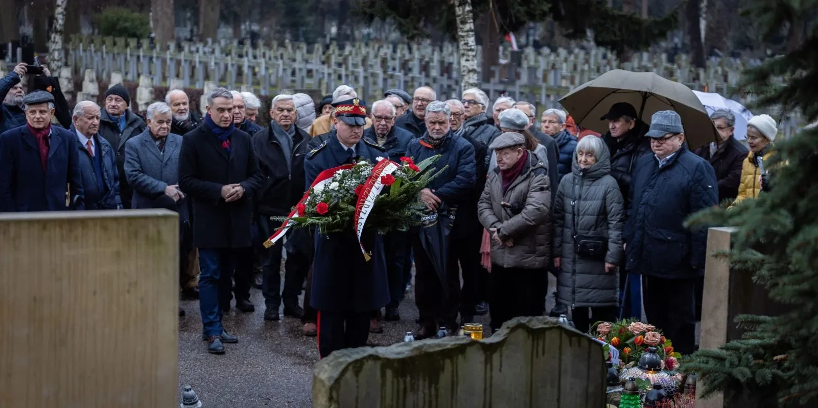 Grupa ludzi na cmentarzu składa wieniec z czerwonych i białych kwiatów w hołdzie pamięci Józefa Oleksyego.