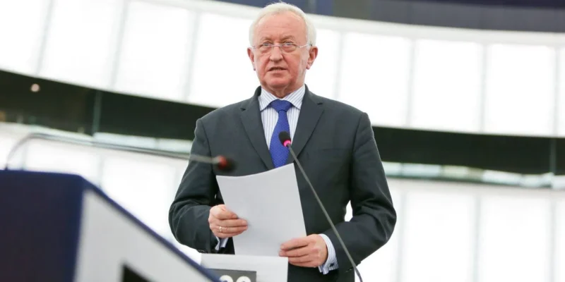 Mężczyzna w garniturze przemawia z kartką w ręku przy mikrofonach na mównicy w sali obrad.