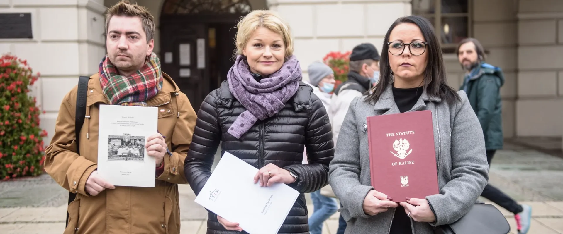 Trzej uczestnicy protestu stoją na tle budynku, trzymając dokumenty i wyrażając swoje zaniepokojenie.