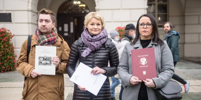 Trzej uczestnicy protestu stoją na tle budynku, trzymając dokumenty i wyrażając swoje zaniepokojenie.