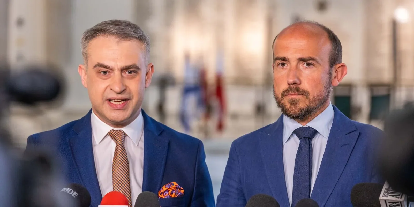 Dwaj mężczyźni w garniturach stoją przed mikrofonami, jeden z nich mówi, a drugi słucha, w tle widoczne flagi.