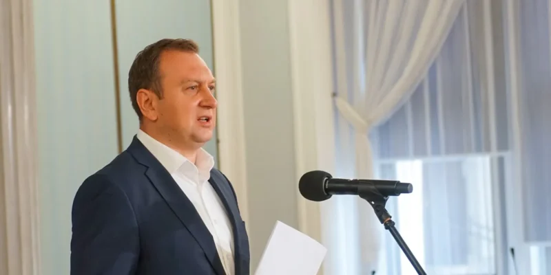 Mężczyzna w garniturze przemawia do mikrofonu, trzyma kartkę z dokumentami, stojąc w jasnym pomieszczeniu z białymi zasłonami.