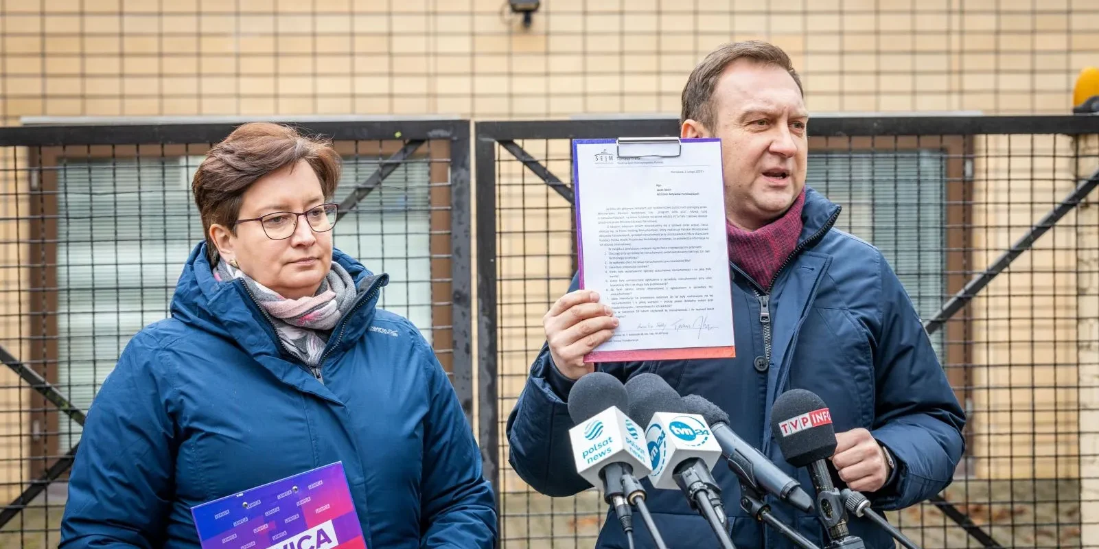 Dwóch polityków Lewicy podczas konferencji prasowej: mężczyzna trzyma kartę z dokumentem przy mikrofonach, kobieta trzyma plakat z napisem LEWICA, stoją przed ogrodzeniem.