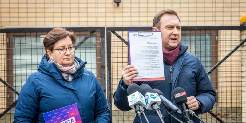 Dwóch polityków Lewicy podczas konferencji prasowej: mężczyzna trzyma kartę z dokumentem przy mikrofonach, kobieta trzyma plakat z napisem LEWICA, stoją przed ogrodzeniem.