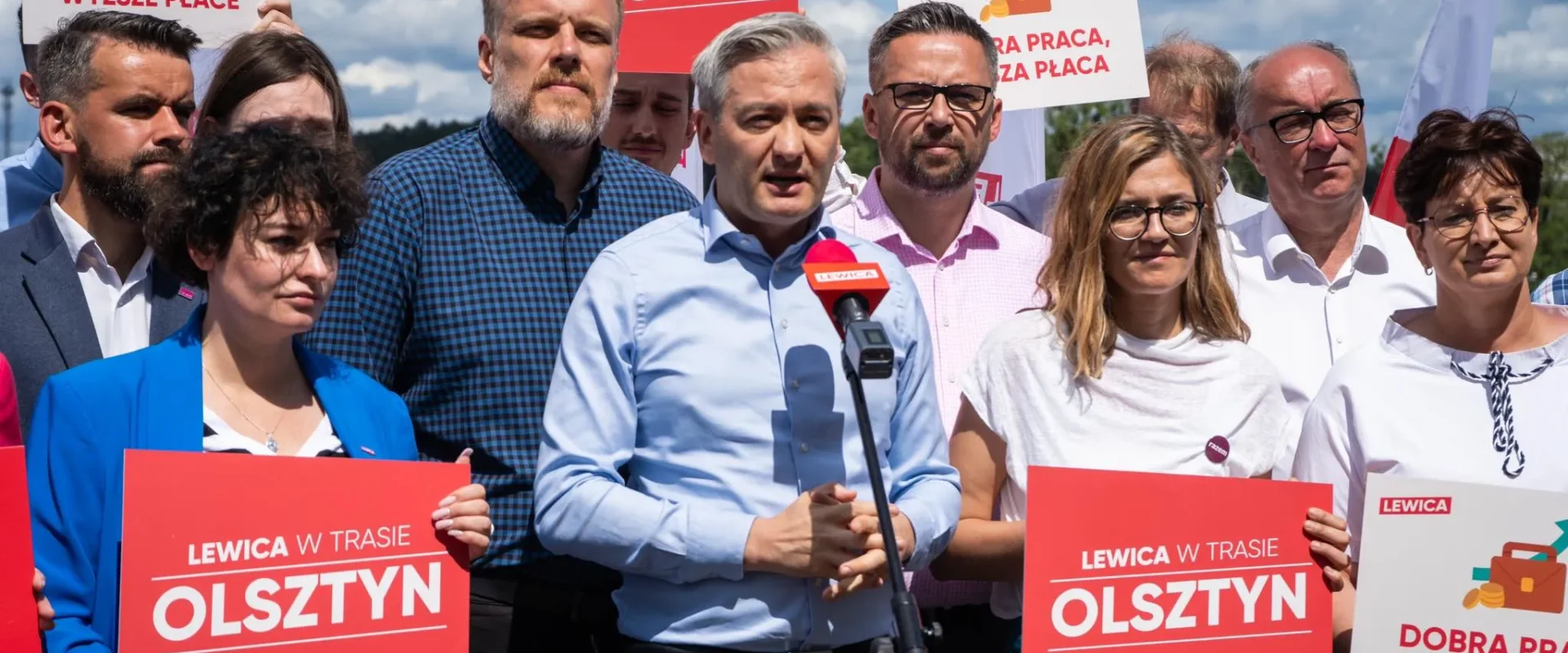 Grupa polityków Lewicy podczas konferencji prasowej w Olsztynie, stojąca za mikrofonem i trzymająca czerwone plakaty z napisem OLSZTYN oraz hasłami wyborczymi.