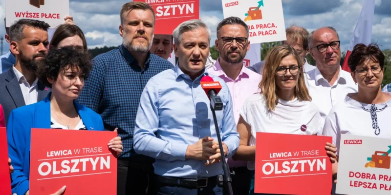 Grupa polityków Lewicy podczas konferencji prasowej w Olsztynie, stojąca za mikrofonem i trzymająca czerwone plakaty z napisem OLSZTYN oraz hasłami wyborczymi.