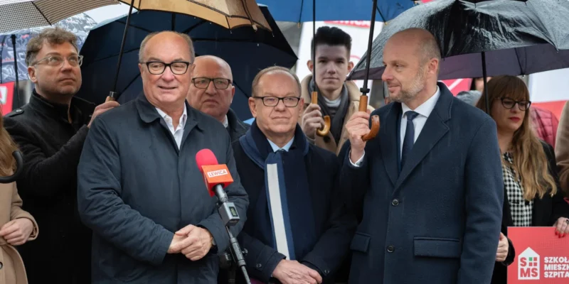 Grupa polityków podczas konferencji prasowej w deszczu, stoją pod parasolami, a przed mikrofonem z logo Lewica przemawia jeden z uczestników.