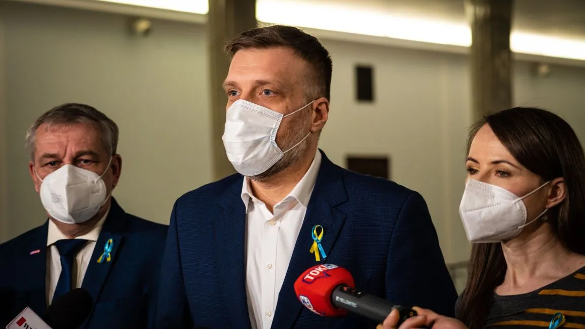 Trzy osoby w maseczkach podczas konferencji prasowej wspierającej uchodźców z Ukrainy, z żółto-niebieskimi wstążkami przypiętymi do ubrań.