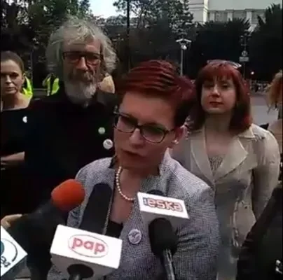 Przedstawicielka Komitetu „Ratujmy Kobiety” udziela wywiadu otoczona mikrofonami i towarzyszącymi osobami na tle budynku.