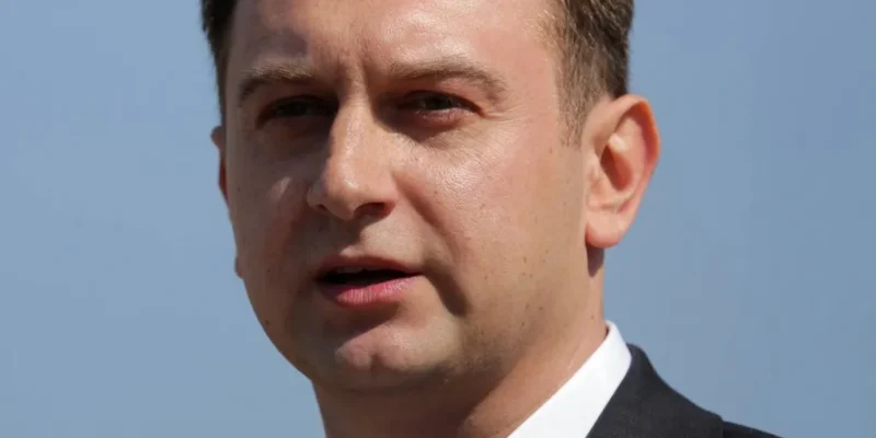 Zbliżenie na mężczyznę w marynarce i białej koszuli przemawiającego na zewnątrz na tle jasnego nieba.