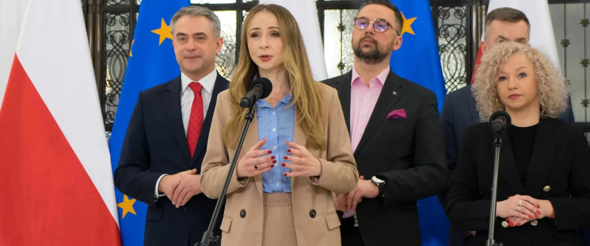 Kobieta w beżowym garniturze przemawia na konferencji prasowej o składce zdrowotnej, stojąc przy mikrofonie, w tle flagi Polski i Unii Europejskiej.