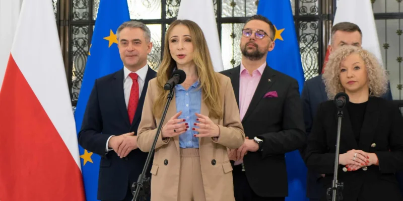 Kobieta w beżowym garniturze przemawia na konferencji prasowej o składce zdrowotnej, stojąc przy mikrofonie, w tle flagi Polski i Unii Europejskiej.