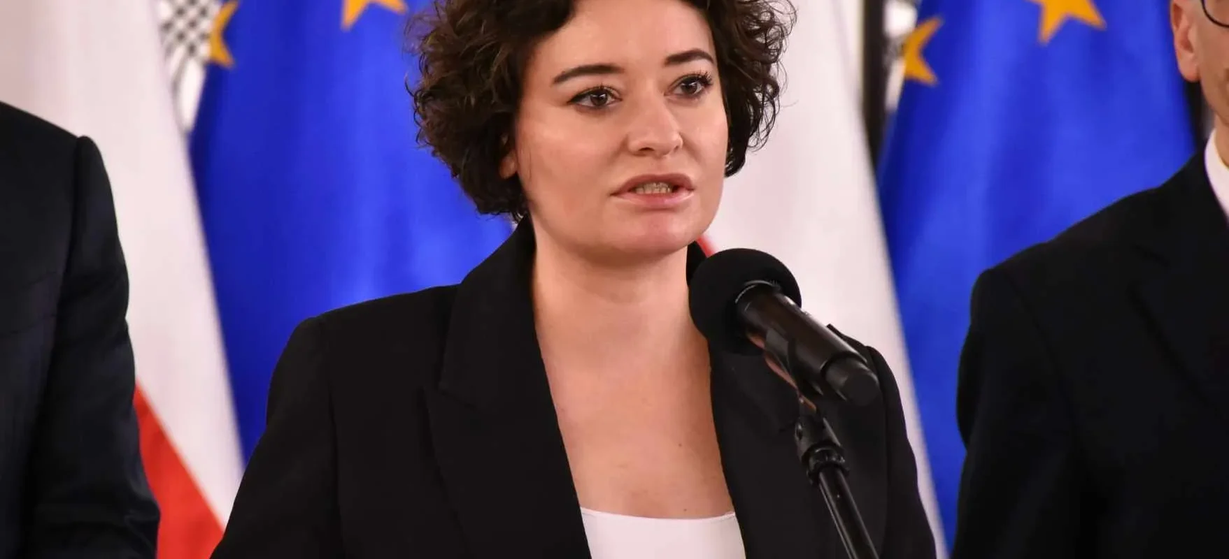 Kobieta w czarnym garniturze przemawia do mikrofonu podczas konferencji prasowej, z flagami Unii Europejskiej w tle.