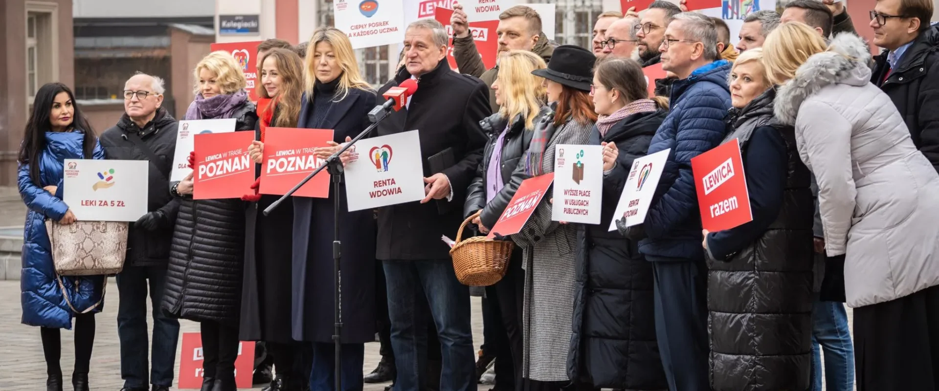 Grupa osób na demonstracji w Poznaniu trzyma plakaty z hasłem Lewica razem, a jeden mężczyzna przemawia do mikrofonu.