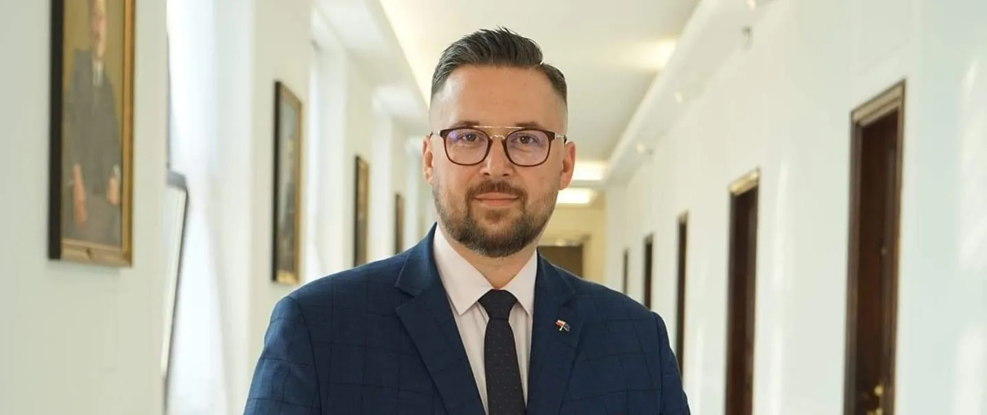 Mężczyzna w granatowym garniturze ze szkłami okularów stoi w jasnym korytarzu z obrazami na ścianach.