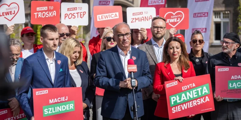 Grupa zwolenników Lewicy podczas plenerowego wiecu, z centralnym mężczyzną w garniturze mówiącym do mikrofonu, wokół którego trzymane są plakaty z nazwiskami kandydatów i logiem Lewicy.