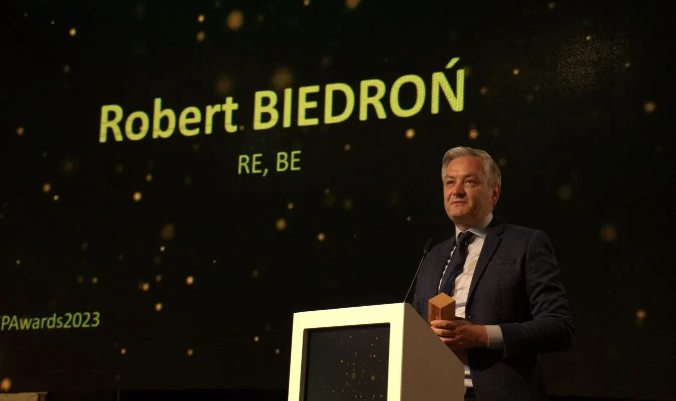 Mężczyzna w garniturze stoi przy podium na scenie podczas ceremonii, a w tle widoczny jest duży ekran z napisem i błyszczącymi punktami świetlnymi.